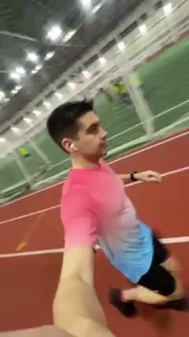 Here's a description of the video:

A man jogs along a red indoor running track while holding a camera. He's wearing a pink and blue gradient t-shirt, black shorts, and wireless earbuds. The track is indoors with bright overhead lighting. There's a green athletic field visible in the background, and bleachers run alongside the track. Other people can be seen on the field in the distance. The camera is shaky due to the man's movement. He occasionally looks into the camera.