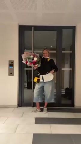 A blonde woman stands in a tiled entryway. She holds a bouquet of flowers in her left hand, while her right hand is slightly raised. She is wearing a black jacket, a white shirt underneath, light blue culottes, and black and white checkered slip-on shoes. She also has a yellow crossbody bag. Behind her are gray glass doors and to her left is an intercom system on the wall. The ceiling is white with a grid pattern.
