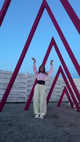 A young woman is performing a dance in an outdoor setting, likely in Pad', Russia, during the evening or dusk hours, indicated by the clear blue sky. She stands on a gravel surface, framed by an art installation consisting of multiple large, pink, triangular wooden structures arranged to form a tunnel or pathway. A white wooden fence is visible in the background.

The woman is dressed in a light pink zipped hoodie worn over a black crop top, paired with light cream or off-white baggy trousers and white shoes. Her dark hair is styled down. She moves fluidly, raising and lowering her arms, swaying her body, and taking small steps, embodying the choreography with focused expressions that suggest confidence and enjoyment. The dance appears expressive and contemporary. The overall scene has a calm yet artistic ambiance due to the soft lighting and the unique architectural elements. No visible text is present in the video.
