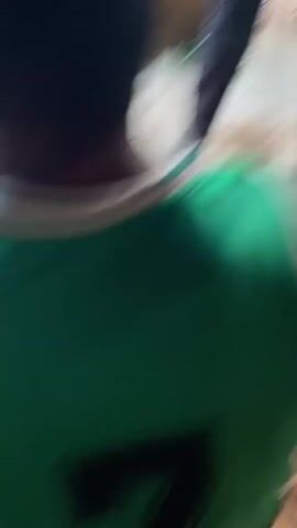 The video features a close-up, low-angle shot of a person from behind, with their back and right arm/shoulder visible. The individual has dark skin and is wearing a vibrant green short-sleeved garment, possibly a t-shirt or sports jersey, which has distinctive white stripes on the shoulder. The fabric appears slightly ruffled.

The person is positioned low to the ground, possibly sitting or crouching, as the camera is angled upwards. The immediate foreground shows a rough, light-colored ground surface, resembling worn concrete or stone, with visible dirt and minor debris. A small, light-colored, cylindrical object, potentially a discarded cigarette butt or piece of rolled paper, is lying on the ground towards the bottom-left of the frame. A dark, blurred object, possibly a shoe or another person's clothing, is partially visible in the bottom-right corner.

The scene appears to be set outdoors or in a semi-enclosed, rustic environment, consistent with the location context of Ogbu, Nigeria. The lighting suggests it is daytime, although specific weather conditions are not discernible. The video is characterized by some camera movement, implying it was recorded handheld. The clip includes background audio where a voice can be heard. No text is visible within the frame. The overall impression is of an candid, unposed moment.