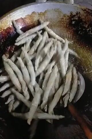 A close-up shot shows many small, thin, light-colored food items being fried in a large, round, metal pan. The food items are stick-shaped and appear to be coated in batter. They are submerged in hot oil, which is bubbling around them. A metal slotted spoon with a wooden handle is used to stir and agitate the food, causing it to move and tumble in the oil. The pan itself is dark and appears to be well-used, with some discoloration and charring on the interior surface. The overall lighting is dim, focusing attention on the cooking process.