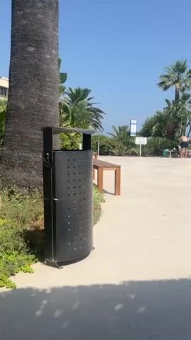 The video pans from a large, dark grey, perforated trash can positioned next to a palm tree trunk, to a blue swimming pool with several people in it. To the right of the pool, there is a white-tiled area with lounge chairs and umbrellas. In the background, there are palm trees, lush greenery, and a building with balconies. The sky is clear and blue, indicating a sunny day.
