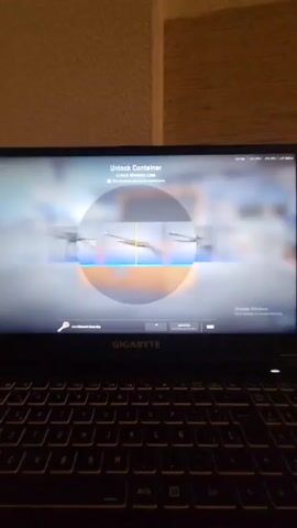The video shows a laptop screen displaying a video game, specifically a container unlock animation from Counter-Strike. The screen reads "Unlock Container, Unlock Kilowatt Case." The animation shows a rotating selection of weapons, and it eventually lands on and displays the "XM1014 | Irezumi" weapon. The laptop is a GIGABYTE brand, with a black keyboard visible below the screen. The background is a neutral-toned wall.