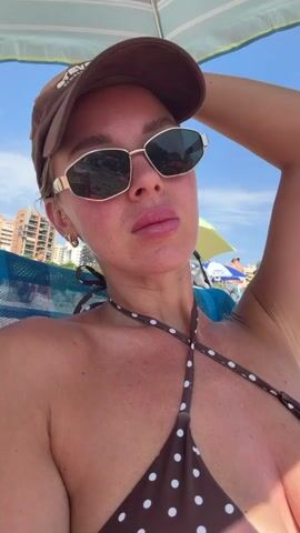 This is a selfie of a woman on a beach. She is wearing a brown polka dot bikini, a brown baseball cap, and sunglasses. She has her arm raised, holding her head. The background shows a beach with lounge chairs, umbrellas, and buildings. The sky is blue and sunny, suggesting it is daytime and good weather. The woman appears relaxed and enjoying the sun. The location context provided is Urbanització Cales I Talaies, Spain. There is some text visible on the baseball cap, but it is too blurry to read.