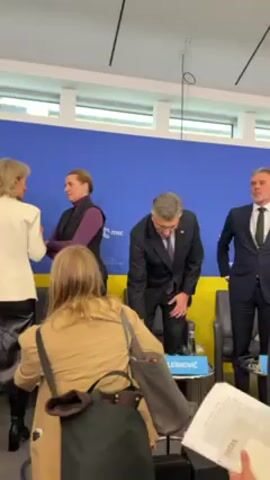 The video shows three individuals, two men in suits and a woman in a vest and shirt, standing in front of a blue backdrop with yellow accents and the logo "msc". The man in the middle is labeled "Plenković" on a sign placed on the table in front of him; the man to his right is labeled "Schoof"; and the woman to his left is labeled "Eriksen". Plenković adjusts his jacket, smiles, and clasps his hands together. Schoof adjusts his tie and looks to the side. People and camera equipment are visible in the foreground, suggesting a press or media event. The room is brightly lit.