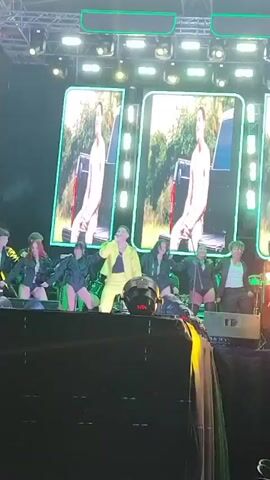 The video shows a live performance on a stage. A man in a bright yellow outfit is in the center, singing and playing a drum.  Several dancers sit behind him, moving to the music.  Two large screens show a close-up of a man singing. The stage is lit with green lights, and there is a crowd visible in the foreground. The overall setting suggests a concert or music festival.