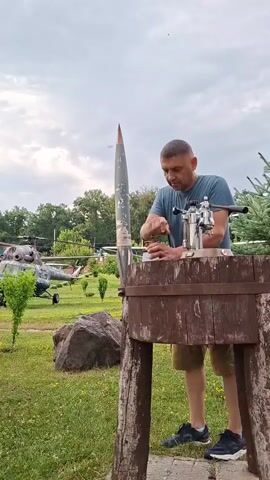 A man stands in a grassy outdoor area, in front of a wooden structure. He is wearing a blue t-shirt and shorts, and is working on a silver object that is attached to a metallic contraption on top of the wooden structure. To his left, a tall, pointed, silver object, resembling a missile, is propped upright against the wooden structure. In the background, two helicopters are visible, along with trees and a cloudy sky.