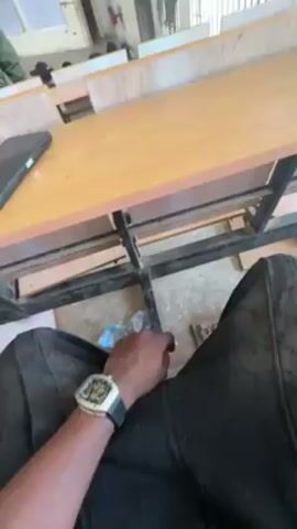 The video shows a person sitting at a wooden desk in a classroom. There are other people in the classroom, and some are walking around. A cell phone is on the desk. The person is wearing a watch with a silver face and black strap.