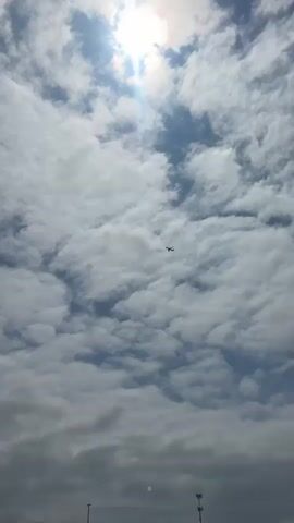 The video shows a bright, partly cloudy sky with the sun visible in the upper portion. A small airplane is seen flying in the distance, silhouetted against the clouds. The clouds are white and puffy, with patches of blue sky visible between them. The bottom of the frame shows the tops of what appear to be light poles. The camera is pointed upwards, capturing the airplane and the sky. The video appears to be a static shot.