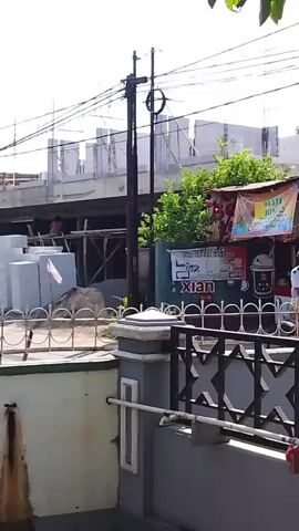 The video begins showing a construction site with buildings under construction and scaffolding. A person is visible on the scaffolding. Several motorcycles are seen on the road in front of the construction site, with some people riding them. Piles of building materials, including bricks and concrete blocks, are stacked at the site. A utility pole with electrical wires stands prominently near the construction. A sign with the word "Xian" is visible on a building adjacent to the construction. A potted plant is also in the foreground, with a fence separating it from the street. The scene appears to be in an urban or suburban environment, with trees and other buildings in the background, under a clear sky.