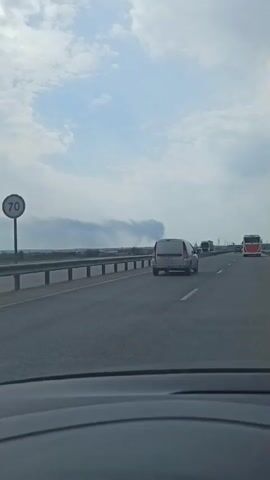 The video shows a view from inside a moving car on a highway. The sky is partly cloudy, and there is a plume of dark smoke visible in the distance, rising from an area beyond the highway. There are several other vehicles on the road, including a silver van, a large truck, and a red and white bus. The car is traveling on the right lane of the highway, and there is a guardrail separating the road from the landscape beyond.