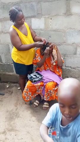 A woman with braided hair is standing behind a seated woman and braiding her hair with a metal tool. The seated woman is wearing a patterned orange outfit. A bag rests on her lap. A young bald child is in the foreground. The scene is outdoors, in front of a cinderblock wall.