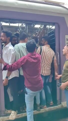 A crowded train car is shown with passengers standing in the doorway and spilling out onto the platform. People are hanging onto the overhead bars and the door frame. The train is purple and white with advertising for "Western Railway" and "ORS Electral" on the side. One person is wearing a white shirt and black pants, another a maroon shirt and blue jeans, and another a striped shirt and blue jeans. The scene appears to be a busy urban environment.