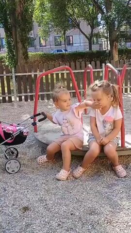 Two young girls are sitting on a merry-go-round in a playground. The girl on the left, wearing a pink t-shirt and shorts, is pushing a stroller with her right hand. She is looking at the girl on the right. The girl on the right, wearing a pink t-shirt and patterned shorts, is covering her face with her left hand. The merry-go-round is made of metal with red supports. The ground is covered in gravel. There is a wooden fence in the background, and trees and buildings are visible beyond the fence. The lighting suggests it is daytime.
