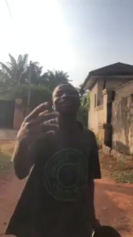 A young man wearing a Versace Jeans Couture t-shirt is standing on a dirt road. He is looking up and smiling. He is gesturing with his hands. There is a building and a fence in the background. The sky is clear and sunny.