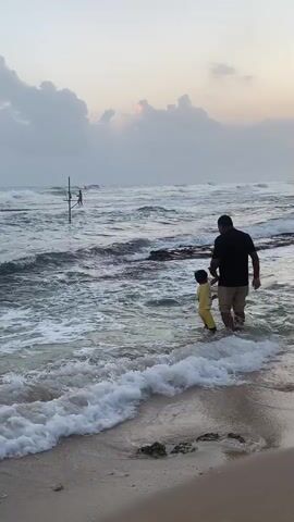 A man and a child are standing in the shallow water at the edge of a beach. The man is wearing a dark t-shirt and light-colored shorts, and the child is wearing a yellow outfit. Waves are gently breaking on the shore. In the distance, a traditional stilt fisherman is visible on a wooden pole in the water. The sky is overcast with clouds, and the sun is peeking through, casting a soft light on the scene. The overall atmosphere is peaceful and serene.