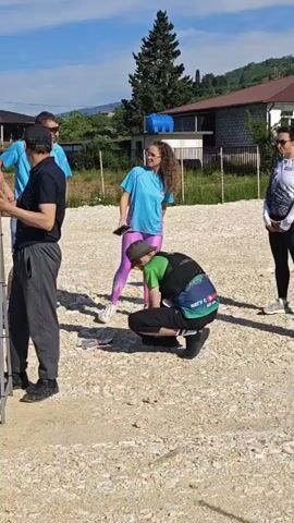 A group of adults stands on a gravel surface outdoors on a sunny day. In the foreground on the left, a man in a black shirt and gray pants stands with his back to the camera. To his right, a man in a green shirt and black shorts faces the camera, holding a small object. Next to him, a woman in a light blue shirt, gray pants, and a black camera around her neck looks at the camera. In the center, a man in a green and blue shirt, black pants, and a hat crouches on the ground, seemingly focused on something below. To his right, a woman in a light blue t-shirt and pink leggings stands with her hands on her hips, looking towards the crouching man. Further right, a woman in a white and black long-sleeved shirt and black pants stands observing the scene. In the background, there are houses, trees, and hills under a clear blue sky.