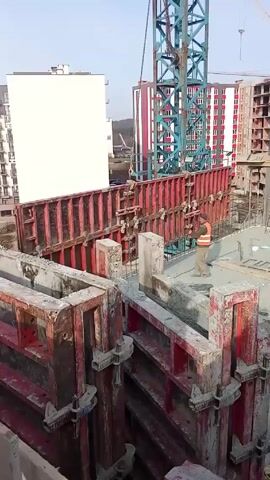 The video shows the construction of a building, featuring a construction worker using a Hilti DX 450 powder-actuated tool to fasten a metal formwork on the upper level of the building. The worker is wearing work gloves and an orange safety vest.  A crane is visible in the background, along with several other building structures. The formwork consists of red metal frames and panels, and is already partially assembled.