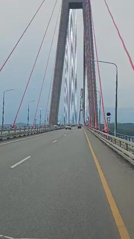 The video shows a view from a vehicle driving on a large cable-stayed bridge. The bridge has two massive concrete towers with red cables supporting the roadway. The roadway is paved and has lane markings. There are streetlights and guardrails along the sides of the bridge. Several cars are visible on the bridge, driving in the same direction as the camera. The sky is overcast. The video has a visual distortion effect applied, causing the bridge towers to appear wavy and distorted.