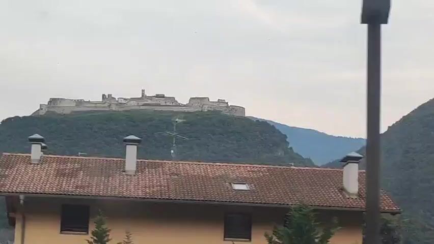 A drone shot slowly pans over a residential building with a red tile roof. The building has two chimneys and several windows. In the background, a large, ancient-looking castle sits atop a verdant, tree-covered mountain. The sky is overcast and a few distant mountains are visible behind the castle.