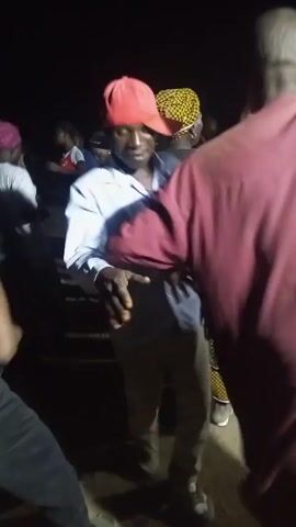 At night, a crowd of people are dancing. A man in a light blue button-down shirt, khaki pants, and a red baseball cap is in the foreground, moving to the music. Other people are around him, some moving to the music and others standing still. Some are wearing headscarves or hats. The lighting is dim.