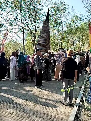A group of people are gathered in an outdoor area with trees and a monument. Some individuals are wearing traditional Islamic clothing. They appear to be participating in an event or procession, as some are carrying items. The monument is tall and appears to be made of stacked wood. The environment is sunny, suggesting daylight.