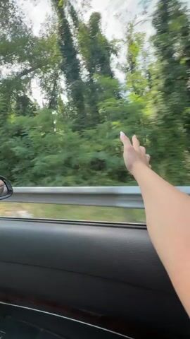 A first-person perspective video shows a person's arm and hand extending out of a car window as the car travels along a tree-lined road. The background consists of lush green trees and foliage. The car's interior, including the door panel and a side mirror, are visible. The person's hand is open, with fingers slightly spread, as if feeling the wind. The video captures the motion of the car, with the scenery blurring slightly in the background.