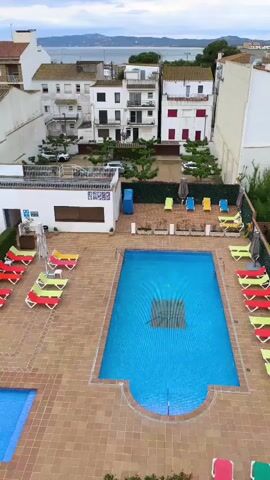 The camera looks down on a scene featuring a rectangular swimming pool filled with blue water, surrounded by a tiled patio area furnished with several lounge chairs in vibrant colors like red, yellow, and green. Behind the pool, a building has a small structure on the roof with a railing and a sign. Further behind the building are several white houses, and in the distance, a large body of water is visible. The sky is overcast with grey clouds, indicating a potentially gloomy or cool day. There are no visible people in the immediate scene. The overall setting appears to be a residential area, possibly a hotel or resort, located near the coast.