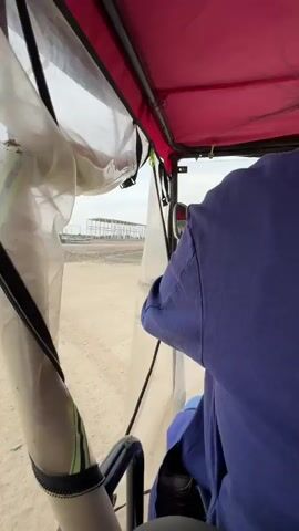 A first-person perspective shot from inside a vehicle, likely a three-wheeled auto-rickshaw, looking out through a transparent plastic enclosure. The vehicle appears to be moving along a dirt or gravel road. Outside, the environment is open and appears to be a construction site or developing area, with some structures and trees visible in the distance under an overcast sky. A person wearing a blue uniform is visible operating the vehicle. There are some insects or debris stuck to the inside of the plastic enclosure. The overall impression is one of transit through a somewhat undeveloped or industrial area.