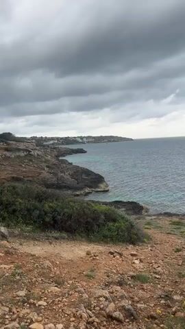 The video shows a rocky coastline on a cloudy day. The camera pans from left to right across the scene, starting with a view of a coastline with buildings in the distance, then moving to a wider shot of the sea. The sky is filled with grey clouds, and the water is a mix of blue and green. The foreground is rocky and covered with sparse vegetation. The wind is strong.