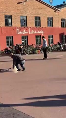 The video shows two people skateboarding in a paved outdoor area. One person wears a cap and blue jeans, while the other wears a white top and black pants. Bicycles are parked in the background near a red brick building with the text "Nørrebro Lokaludvalg" on it. A café sign is visible to the left. One skateboarder attempts a trick and lands on the ground, but continues skateboarding afterwards. The other skateboarder is seen maintaining balance while riding.