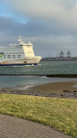 A large white ferry with the words "Stena Line" and "Connecting Europe" on its side is sailing through choppy water. The ferry is white with blue and green accents and a red funnel. The water is a greenish-blue with whitecaps. In the background, there are cranes and industrial buildings, suggesting a port or harbor. In the foreground, there is a strip of grass next to a paved pathway. The sky is cloudy with patches of blue.
