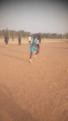 The video shows multiple young men doing agility drills on a dirt field, running in between orange cones set in a row. The players are wearing different colored shirts and shorts. In the background are other players standing around, as well as a soccer goal. The sky is a clear, pale blue.