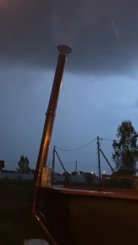The video shows a dark, cloudy sky over a wooden, A-frame structure with lights. Initially, a tall metal pipe is visible, likely a chimney for a stove or fireplace. The camera then pans right to reveal the illuminated A-frame house, decorated with string lights. The string lights extend beyond the house, hanging from wooden posts. The ground appears to be gravel. The general impression is of a cozy, lit-up space against the backdrop of an approaching storm.