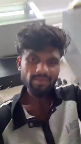 A close-up video shows a man with dark, wavy hair and a beard looking directly at the camera. He is wearing a black and white patterned jacket over a light-colored shirt. The background is a light-colored wall with an electrical socket and wires visible. A poster with "प्लेक्स" (plex) written at the top in Marathi script, along with a calendar-like grid of numbers (१ to ३१) also in Marathi, is visible behind him. The man initially smiles subtly, then runs his hand through his hair, adjusting it, while continuing to look at the camera. His expression remains calm and slightly smiling throughout the short clip. The setting appears to be indoors, possibly an office or shop in Palghar, India, based on the visible script and provided context.