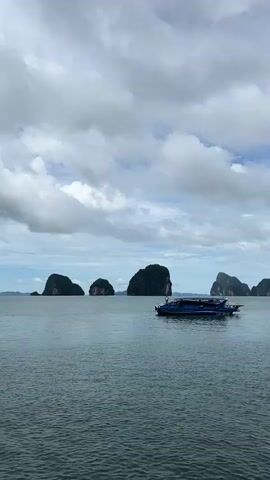A high-angle, vertical shot shows a blue speedboat in the middle of a wide expanse of water, with several rocky islands dotting the horizon. The sky is filled with dramatic, grey and white clouds, suggesting it might be a bit overcast or stormy. The water is a muted blue-grey, with a slight chop. The islands are covered in lush green vegetation, and some have sheer, vertical cliffs. The boat appears to be moving slowly, and there are no people visible on deck, though there may be people inside the cabin. The overall impression is one of a serene but potentially wild natural landscape.