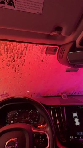 Inside a car at a carwash, the view through the windshield is obscured by soapy water and cleaning brushes. The interior shows a Volvo dashboard, including the steering wheel, speedometer, and a central console screen displaying a car graphic. The lighting in the carwash cycles through various colors.
