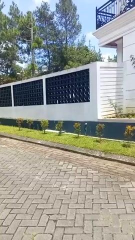 The video shows a camera panning across a residential street paved with interlocking bricks. On the left, a low white wall with decorative black lattice panels runs alongside the road. Behind the wall, there's a grassy area and a line of tall pine trees against a blue sky with scattered white clouds. As the camera pans right, the scene shifts to a view of houses behind a concrete wall. Palm trees are dotted along the landscape. The final shot shows a multi-story house under construction. A street lamp is visible in the foreground.