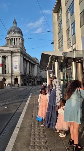 The video opens with a close-up shot of a street scene. A tram stop shelter with a modern glass and metal design is visible on the right side of the frame. A group of people, including several children, are standing at the tram stop, waiting. The children are dressed in colorful, festive clothing, suggesting a special occasion. A woman in a blue dress and headscarf is holding one of the children. The background features an imposing building with a dome and a clock, likely a historical landmark. A large English flag is draped across the side of this building, along with banners that say "Come On England," indicating a sporting event or celebration. The street is paved with tram tracks, and a few cars and pedestrians are visible in the distance. The sun is shining, casting long shadows and creating a warm, golden light, suggesting it is late afternoon or early evening. The sky is blue with scattered white clouds. The overall atmosphere appears to be one of anticipation and community gathering.