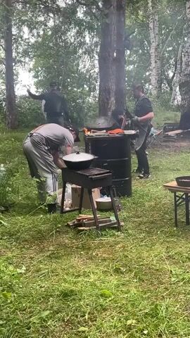 The video shows a group of people cooking outdoors in a grassy, wooded area. In the foreground, a person in a grey apron and grey pants is bending over a grill, tending to a pot. To their right, a black metal barrel is set up with a fire inside, and a person in a black shirt and apron is cooking over it. Smoke is rising from the barrel. In the background, two other people are visible, one wearing a black jacket and the other in a black t-shirt. There is a wooden table to the right of the barrel, with a black pan on it. The environment appears to be a forest clearing with trees and undergrowth. The overall scene suggests a casual outdoor cooking event.