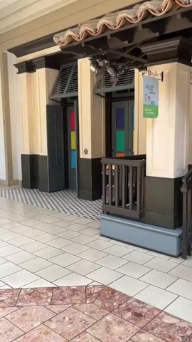 The video shows a small library with bookshelves filled with books, two chairs, and a small table. The library is decorated with striped wallpaper and has a tiled floor. The video also shows a sign that says "A Little Library." The library appears to be located in a public space, possibly a mall or a shopping center, given the tiled walkway and the overhead structure. The lighting is artificial and seems to be coming from the shelves and overhead fixtures. The video is shot from a slightly low angle, looking towards the library entrance.