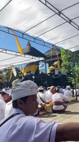 The video shows a gathering of people, mostly men, sitting on the ground outdoors under a white canopy. They are dressed in white shirts and head coverings, suggesting a religious or ceremonial event. In the background, a multi-tiered dark structure, possibly a temple or altar, is visible, adorned with yellow and white banners. The atmosphere appears calm and attentive, with people facing towards the structure. The ground is paved with stone tiles, and the sky is clear blue. The camera is positioned at a low angle, focusing on the people in the foreground and the ceremonial site in the background.