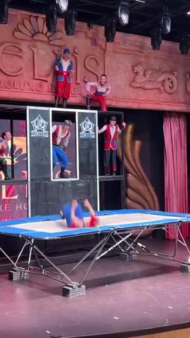 The video shows a performance on a stage. In the foreground, a blue and silver trampoline is set up. Behind the trampoline, there is a multi-level structure with several performers dressed as pirates. On the top level, three performers stand, and one is in mid-air, performing a somersault. On a lower level, two performers are visible, one in a black box and another standing. To the left, another performer in a pirate costume is seen. The background features a large, textured wall with the words "DELUXE HOTEL" visible, and red curtains on the right side. Spotlights are positioned above the stage. The lighting is dramatic, with red and pink hues dominating the scene.