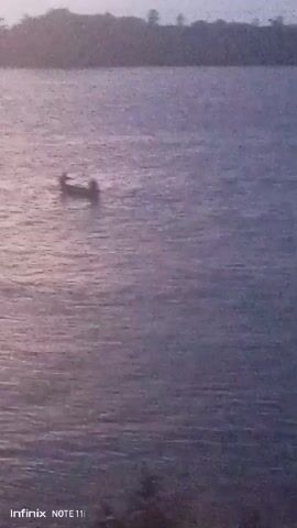 The video shows a view of a body of water with a small boat in the distance. The boat appears to have at least one person in it. The water is calm and slightly reflective. There is vegetation visible on the far shore. The scene is filmed in the late afternoon or early morning light. The quality of the video is somewhat grainy. The lower left corner of the frame has "Infinix Note 11i" on it.
