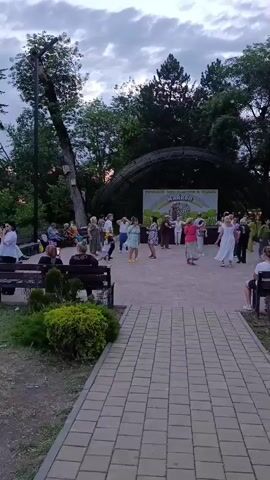 An outdoor gathering is taking place in a park-like setting. Several people, predominantly older adults, are dancing in a circular formation in front of a stage. The stage has a large banner with colorful text and imagery. Other individuals are seated on benches around the dancing area, observing the event. Trees and greenery surround the open space, and a paved pathway leads towards the gathering. The lighting suggests it is late afternoon or early evening, with a cloudy sky overhead.