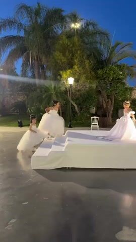 The video captures an outdoor event at dusk, likely a wedding or a formal ceremony, in the Beer Tuvia Regional Council, Israel. The scene is set against a backdrop of palm trees and lush greenery, illuminated by ambient lighting. A raised white platform, draped in fabric, serves as the central stage or runway.

Several young girls, dressed in elaborate white dresses that resemble communion or flower girl attire, are present. One girl in a particularly voluminous dress is positioned on the platform, with another younger girl in a similar dress near the base of the platform. Further down the platform, another girl in a white dress is visible, possibly participating in a procession or performance.

In the background, a stone wall suggests a rustic or historic architectural element. Rows of white chairs are set up on either side of the platform, indicating an audience is present or expected. A few adults are seated in the foreground, facing the platform, and others are standing further back near the stone wall.

The time of day is indicated by the deep blue twilight sky. The overall atmosphere appears to be festive and ceremonial, with a sense of anticipation or celebration. The lighting is soft, highlighting the subjects and creating a warm glow on the stage. The presence of palm trees and the stone wall strongly suggest a Mediterranean or Middle Eastern location, consistent with the specified region. No visible text is present in the video.