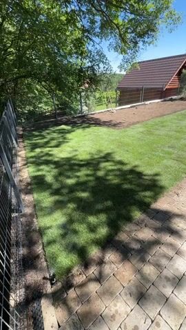 The video shows a newly landscaped yard with a sprinkler system irrigating the fresh grass. A person in shorts and a hat stands on a paver walkway, looking at some equipment in the yard. A small house with a dark roof can be seen in the background, and there is a fence visible along the property line. Tree branches cast shadows on the lawn. The sky is clear and blue.