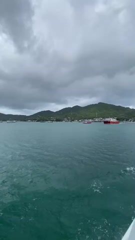 The scene is a wide shot of a body of water with a landmass in the background, under a heavily clouded, overcast sky. The water is a dark teal color with subtle ripples and wakes in the foreground, suggesting the movement of a vessel. Across the water, a series of low green hills or small mountains stretch horizontally. Buildings are sparsely scattered along the coastline, appearing as small white or light-colored structures. A red and white ferry is visible near the shore. The scene is static, captured from a boat, providing a perspective of approaching the land.