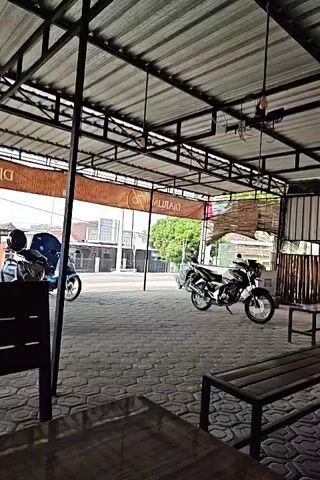The video pans across an outdoor seating area with a corrugated metal roof supported by metal beams. In the foreground, a dark wooden bench is visible. Beyond the bench, a paved area with hexagonal stones is visible. Several motorcycles are parked on the paved area. On the left, two motorcycles are parked closely together, one with a blue windshield. Further back and to the right, another motorcycle is parked. In the background, a building with an orange banner displaying text is visible, along with other buildings and trees. The overall impression is of an open-air, informal establishment, possibly a cafe or rest stop, with parking for motorcycles.