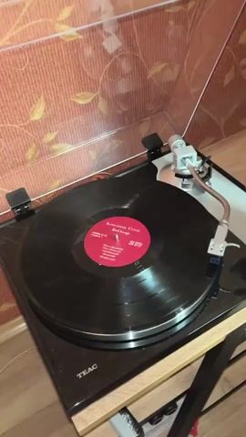 The video shows a close-up shot of a record player with a vinyl record spinning. A person's hand is visible in the top right corner of the frame, gently touching the tonearm. The record has a red label with text that is partially legible. The background appears to be a wall with textured wallpaper. The record player itself is black with a clear dust cover. The overall impression is one of someone preparing to play or adjust a record.